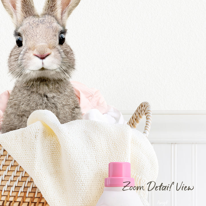 A cute gray rabbit sits in a woven basket filled with clothes, with a pink bottle of laundry detergent placed next to it.