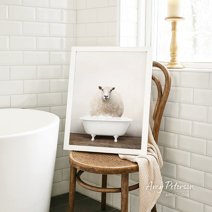 A framed image of a sheep sitting in a bathtub is displayed on a wooden chair in a bathroom setting.