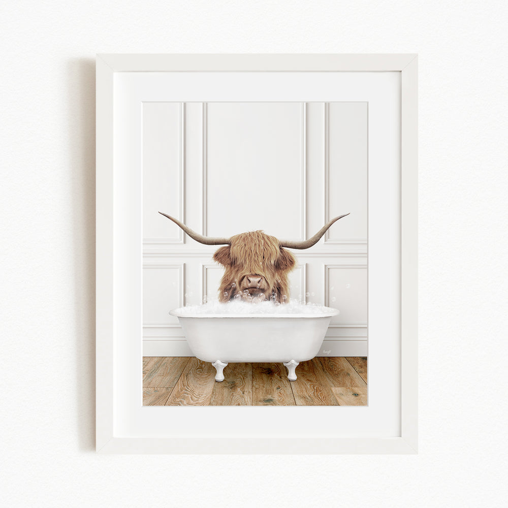 A white framed picture depicting a Highland cow with large curved horns sitting in a white bathtub filled with water.