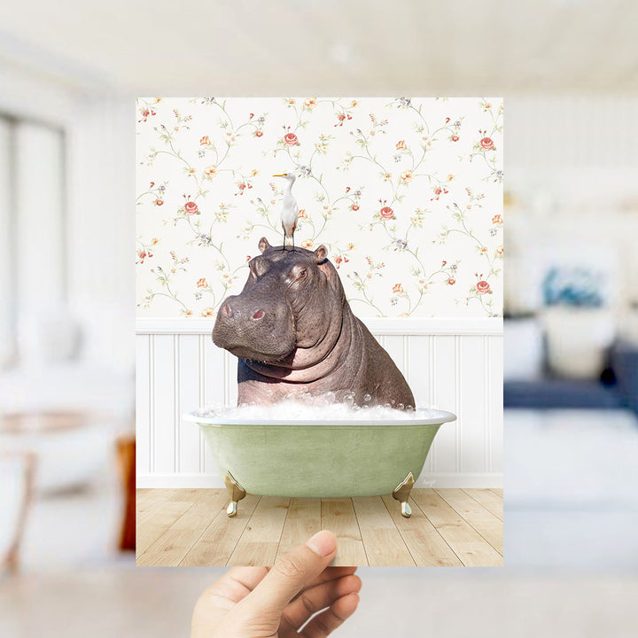 A hand holds up a framed image of a hippopotamus sitting in a bathtub, surrounded by a floral wallpaper.
