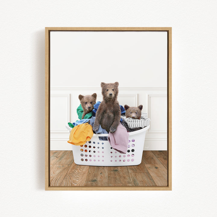 A framed image showing three brown bears sitting in a laundry basket filled with clothes.