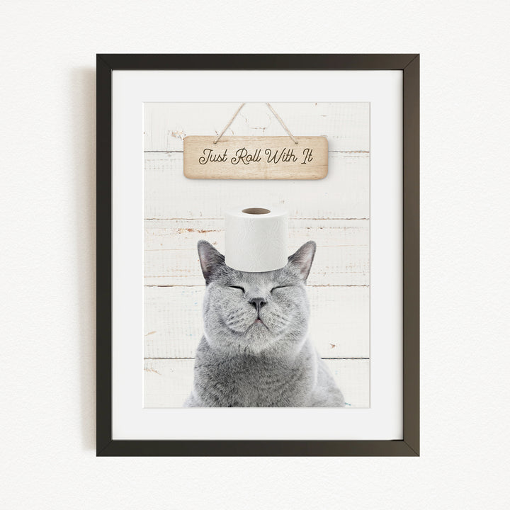 A framed image of a gray cat with its eyes closed, wearing a roll of toilet paper on its head, and a wooden sign above it that says "Just Roll With It".