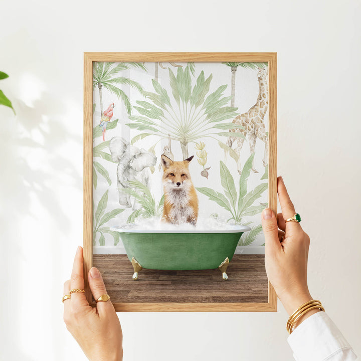 A person's hands are holding a framed picture of a fox sitting in a green bathtub, surrounded by tropical foliage.