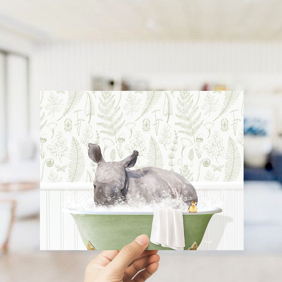 A hand holding a framed image of a rhinoceros sitting in a bathtub filled with bubbles, surrounded by a green and white floral pattern.