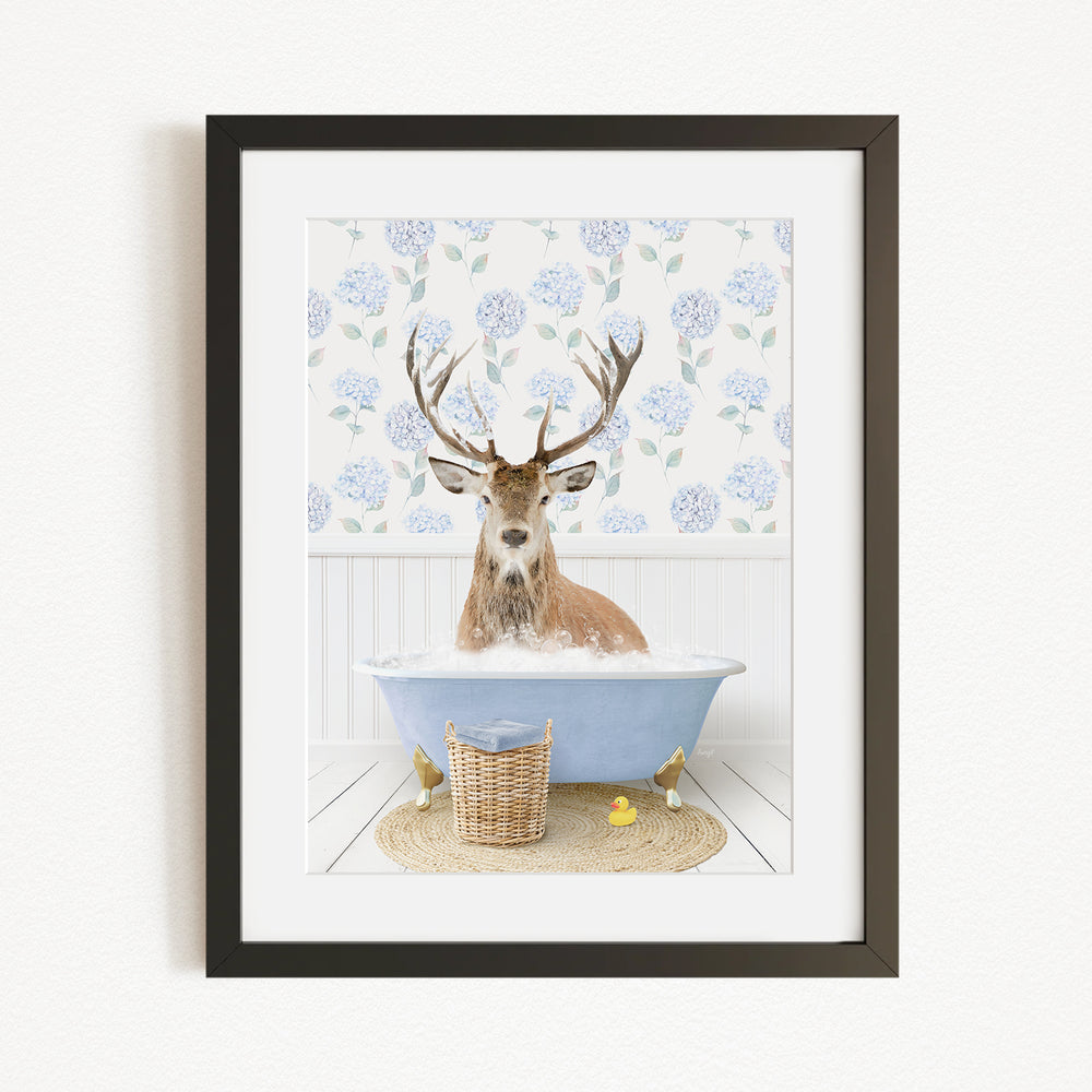 A deer with antlers is sitting in a blue bathtub filled with bubbles, surrounded by a wicker basket and yellow rubber ducks.