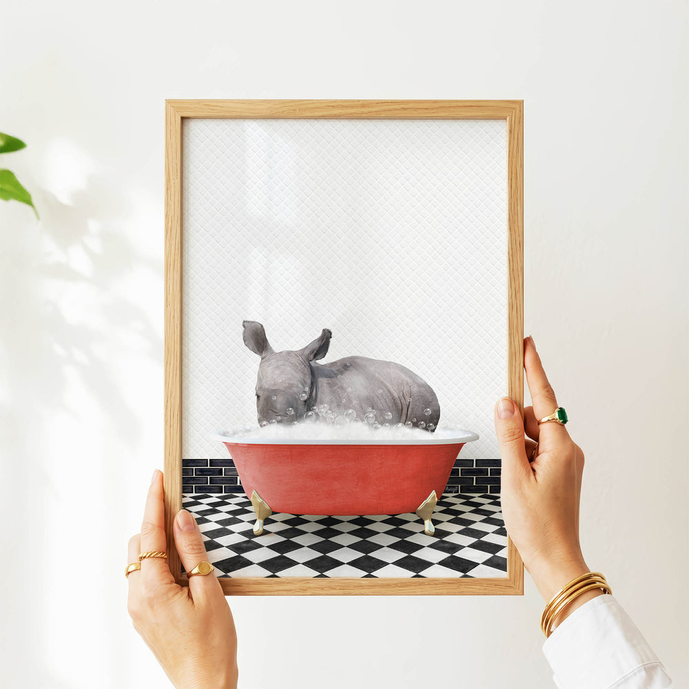 A person's hands are holding a framed picture of a rhinoceros sitting in a red bathtub filled with water, surrounded by a black and white checkered floor.