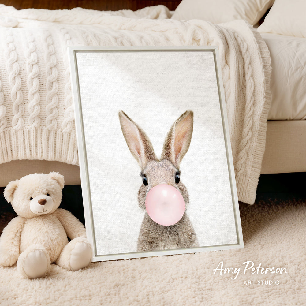A framed image of a rabbit blowing a bubble with a pink bubble gum bubble is displayed on a bed, accompanied by a teddy bear.