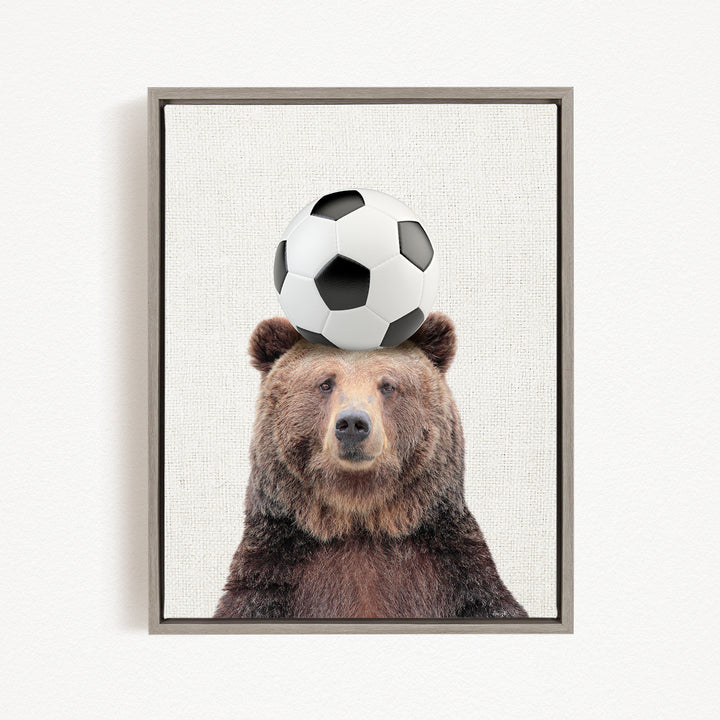 A brown bear with a soccer ball on its head, looking directly at the camera.