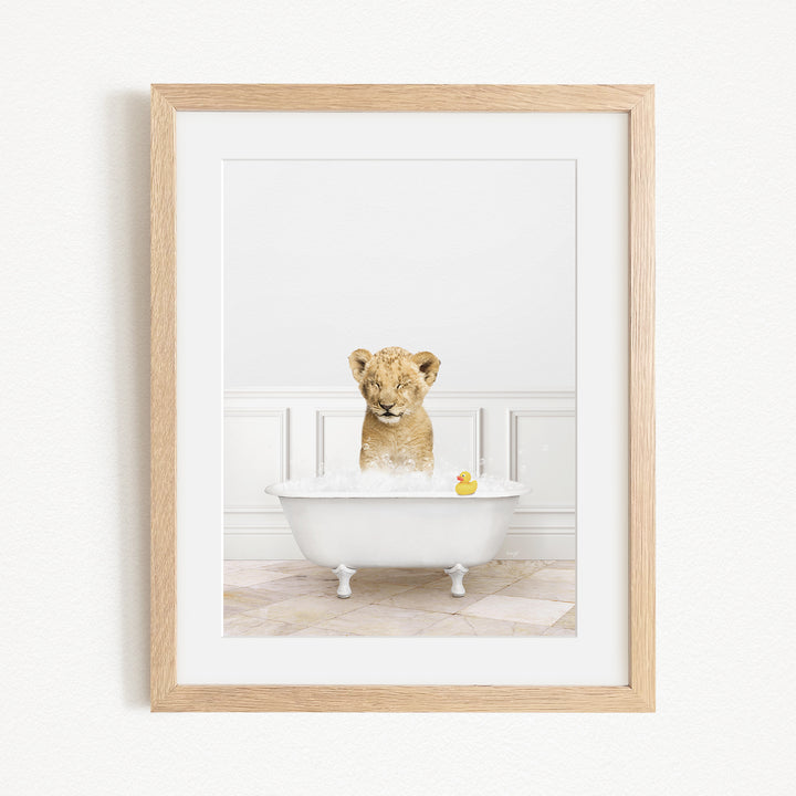 A young lion cub sitting in a white bathtub filled with water and bubbles, with a yellow rubber duck floating in the water.