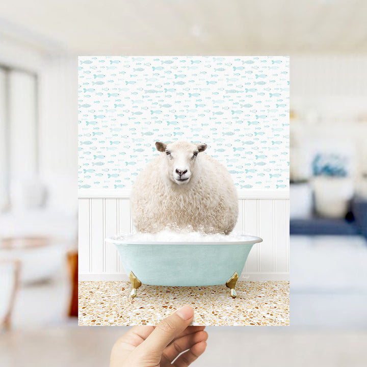 A hand holds up a framed image of a sheep sitting in a bathtub filled with bubbles, surrounded by a pattern of fish.