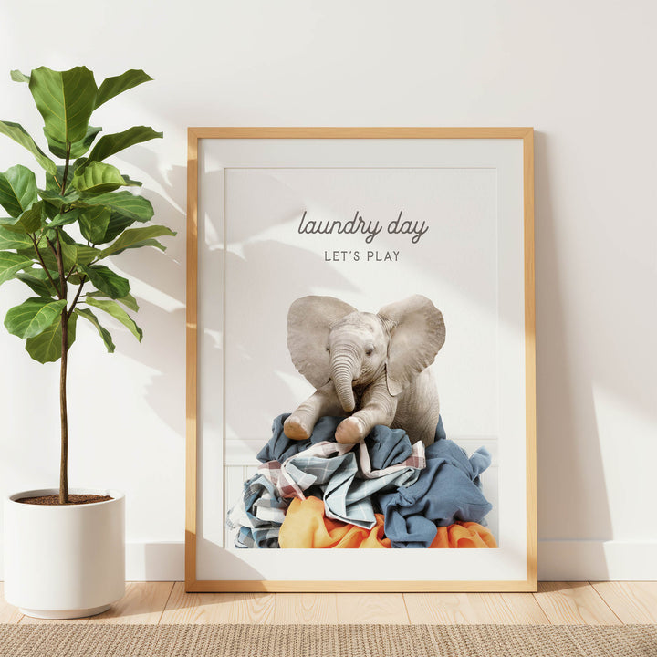 A framed poster with an elephant sitting on a pile of clothes, accompanied by the text "laundry day let's play".