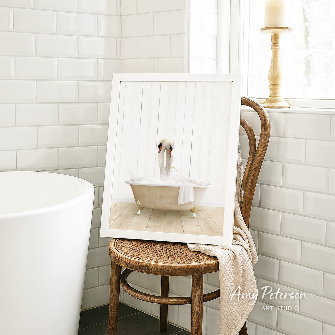 A framed image of a swan sitting in a bathtub is displayed on a wooden chair in a bathroom setting.
