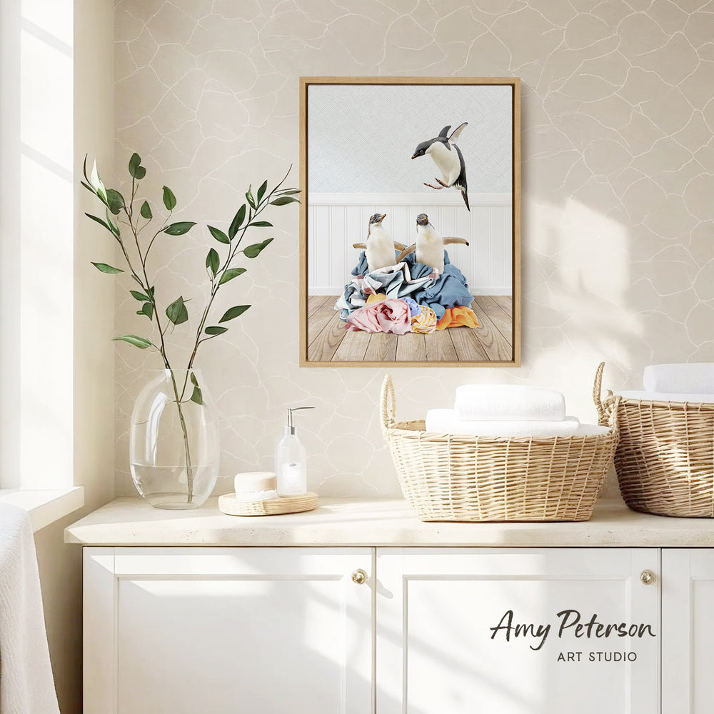 a framed photograph of two penguins on a wooden floor, surrounded by a pile of clothes, with a wicker basket and a vase of leaves nearby.