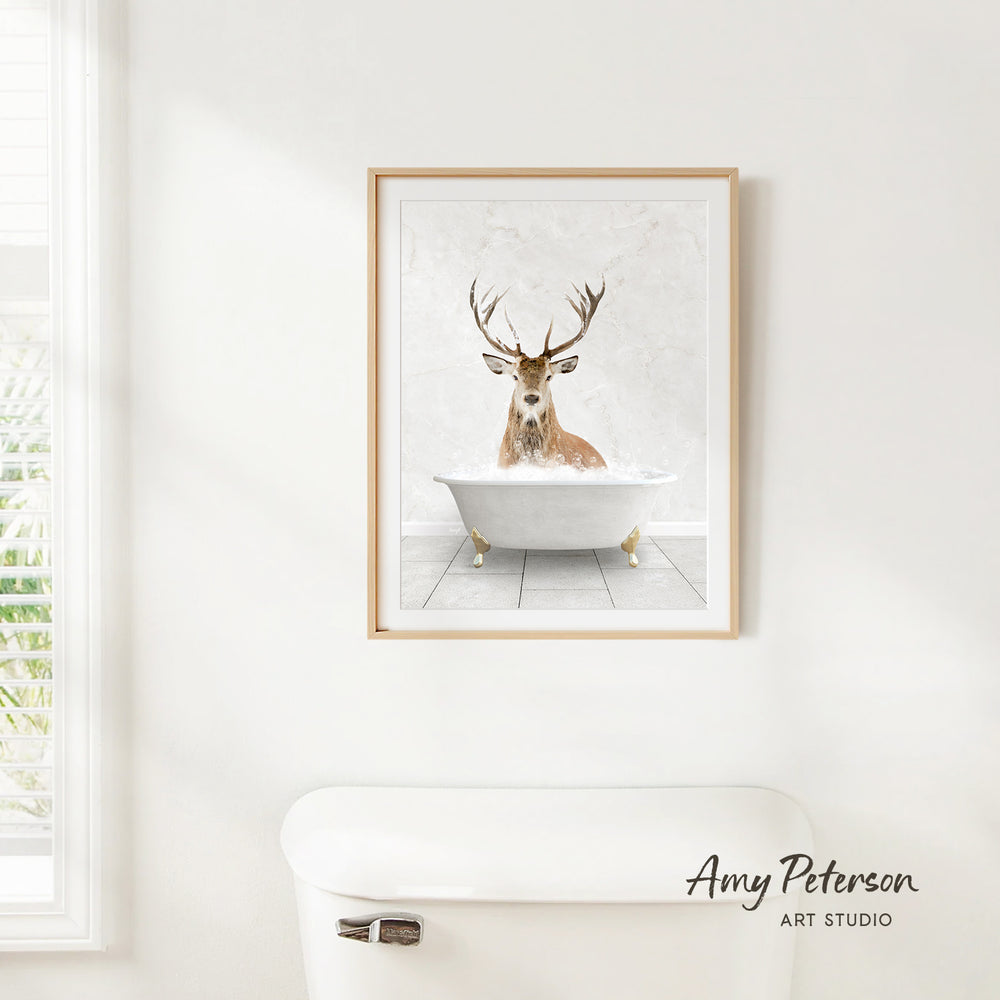 A framed image of a deer sitting in a bathtub, with its antlers raised, is hanging on a white wall.