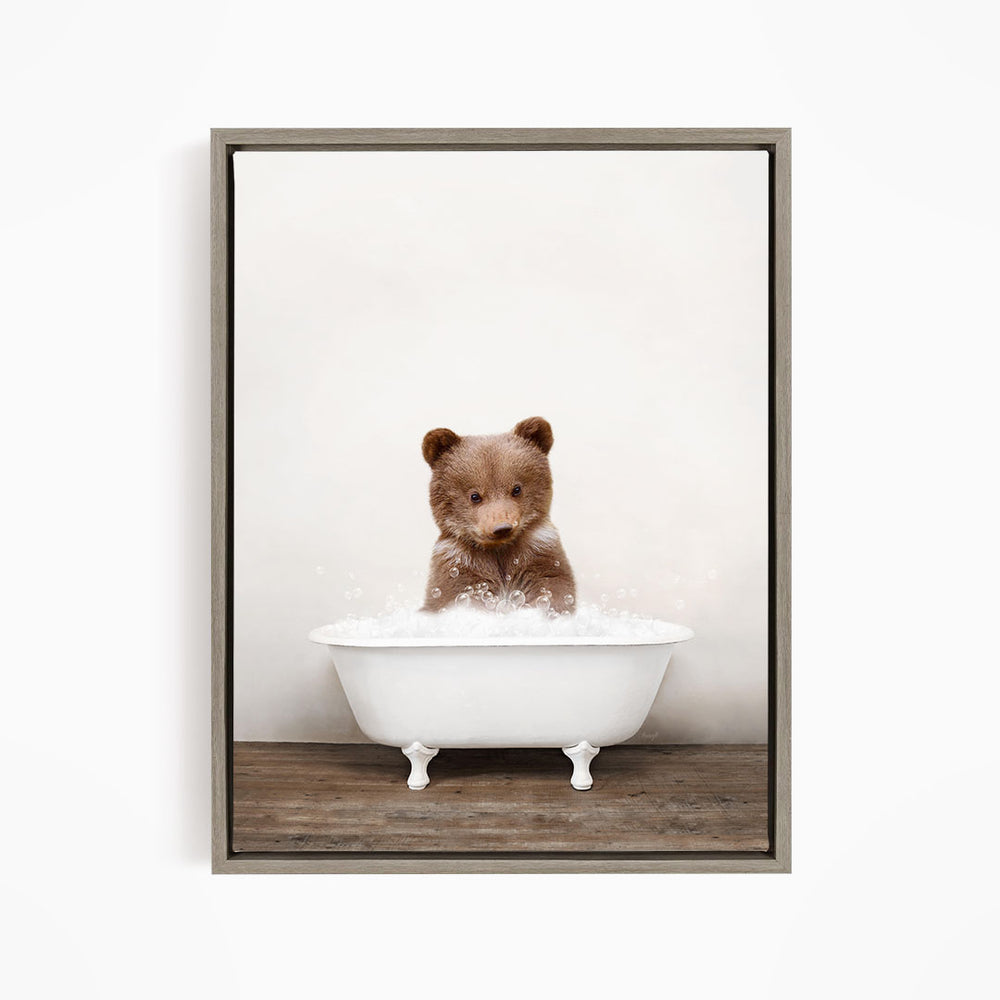 A cute brown bear sitting in a white bathtub filled with bubbles, looking directly at the camera.