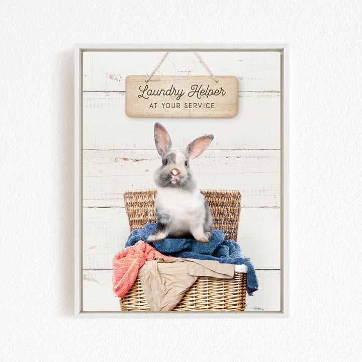 A gray and white rabbit sits in a wicker basket filled with laundry, with a wooden sign above that reads "Laundry Helper at your service".