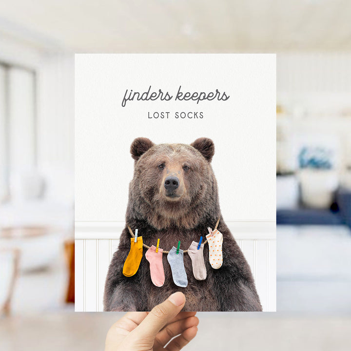 A hand holding a printed image of a bear wearing socks, with the text "finders keepers" and "lost socks" printed above and below the bear.