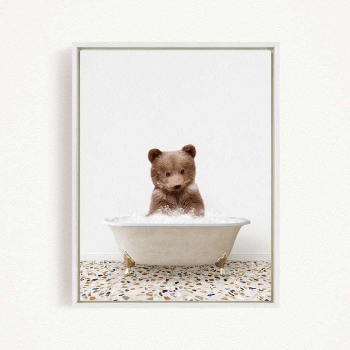 A cute brown bear cub sitting in a white bathtub filled with bubbles, looking directly at the camera.