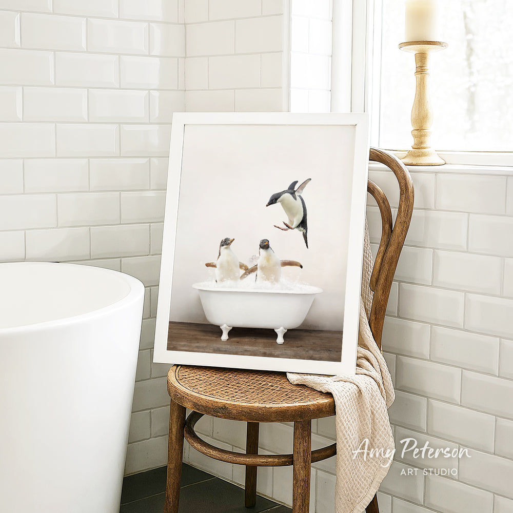 A framed photograph of two penguins sitting in a bathtub, with one penguin standing on its hind legs, is displayed on a wooden chair in a bathroom setting.