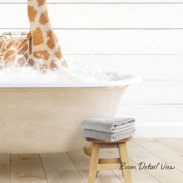 A giraffe standing in a bathtub filled with bubbles, with a towel and stool nearby.