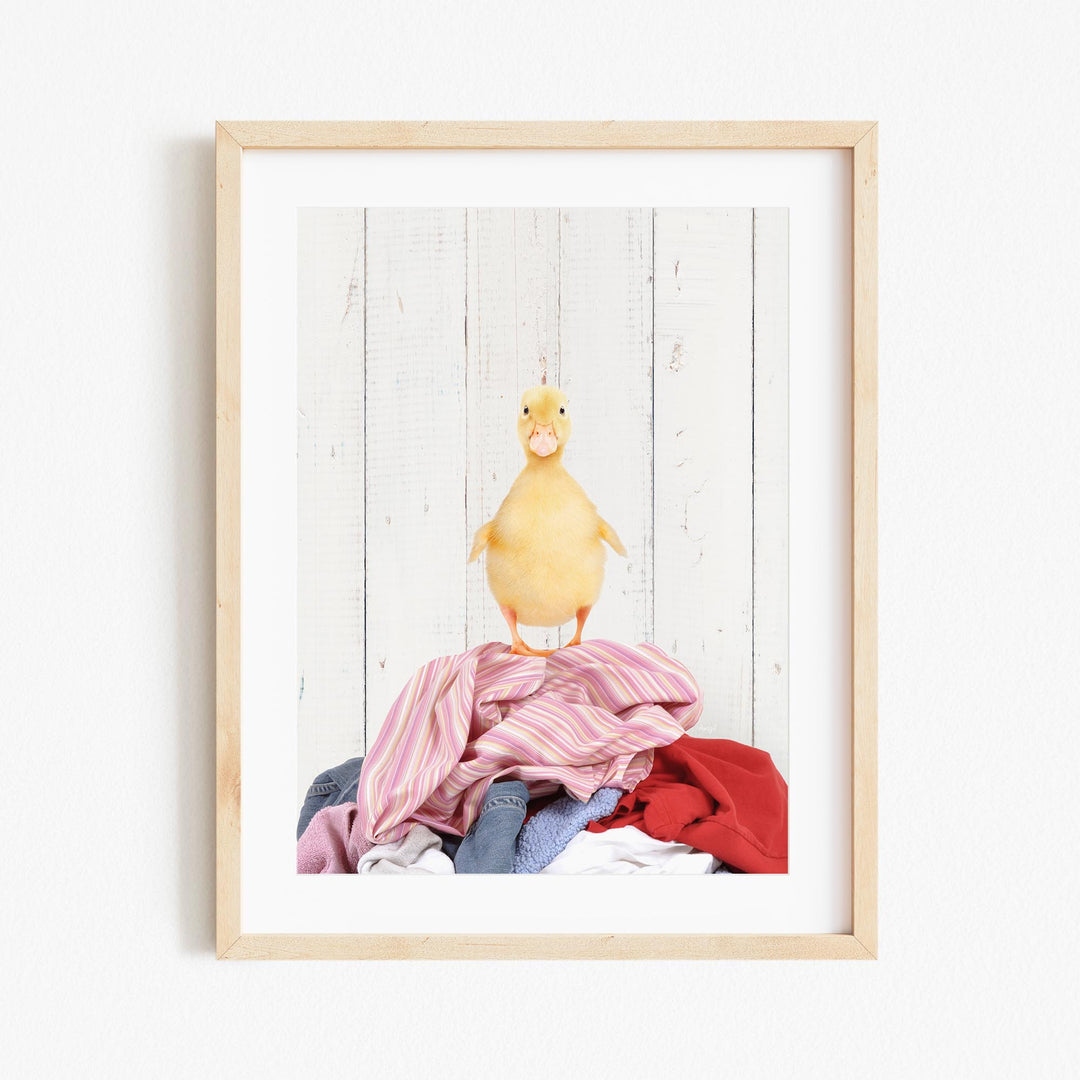 A yellow duck standing on a pile of clothes against a white wooden background.