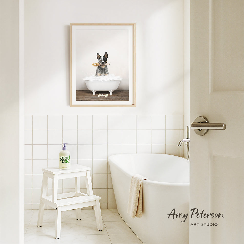 A framed image of a dog sitting in a bathtub, with a towel draped over the side of the tub, is displayed on the wall above a white bathtub in a clean, white-tiled bathroom.