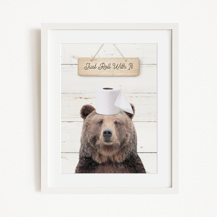 A framed image of a brown bear wearing a toilet paper hat, with a wooden sign above that says "Just Roll With It".