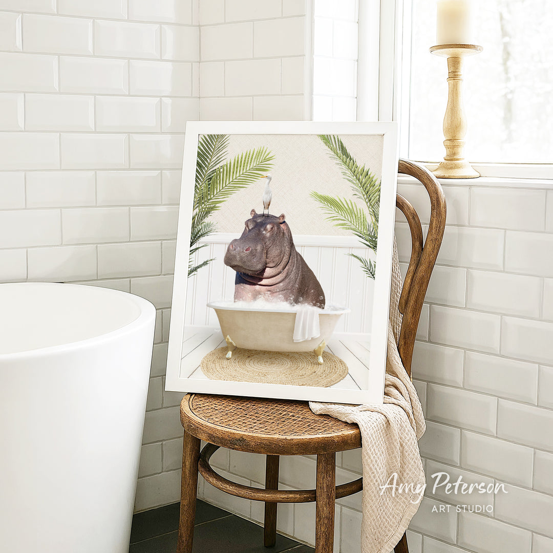 A framed image of a hippopotamus sitting in a bathtub, surrounded by palm leaves, is displayed on a wooden chair in a bathroom setting.