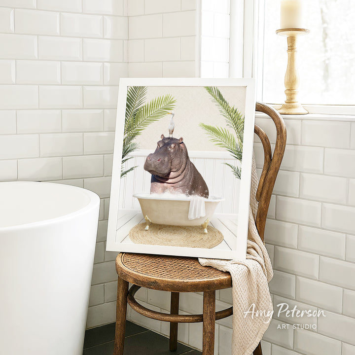 A framed image of a hippopotamus sitting in a bathtub, surrounded by palm leaves, is displayed on a wooden chair in a bathroom setting.