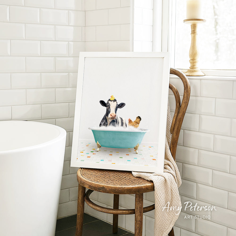 A framed image of a cow sitting in a bathtub, with a chicken nearby, is displayed on a wooden chair in a bathroom setting.