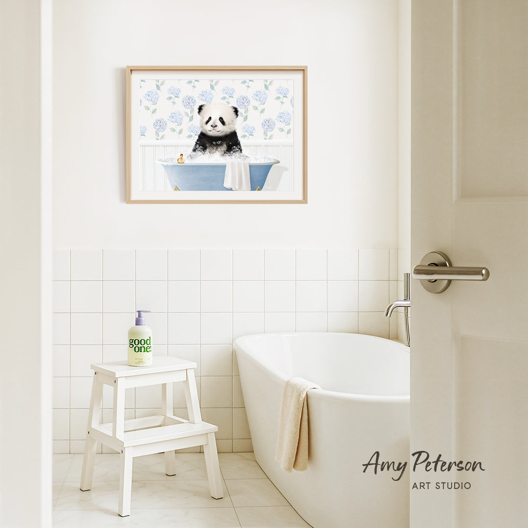 A framed image of a panda sitting in a bathtub is hanging on a white tiled wall above a white bathtub in a bathroom.