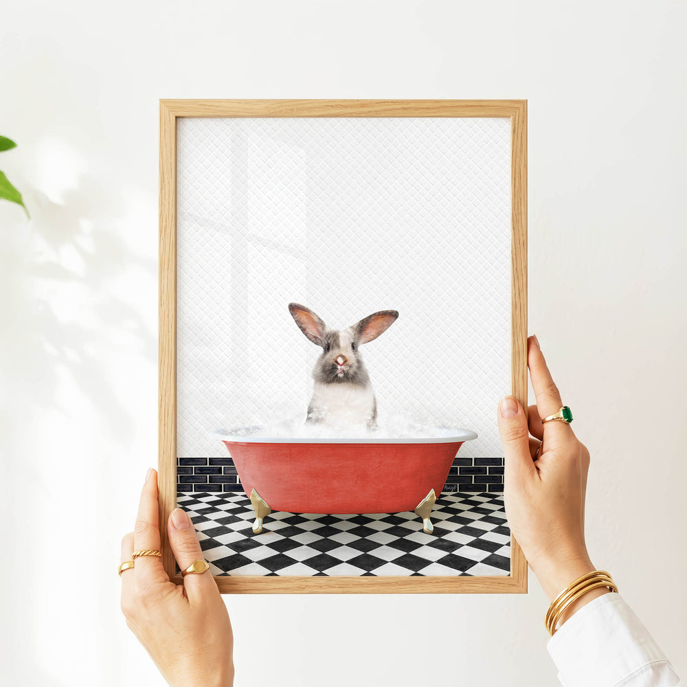 A person's hands are holding a framed picture of a rabbit sitting in a red bathtub filled with water, with a black and white checkered floor visible beneath the tub.