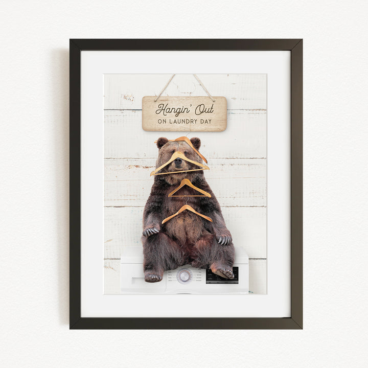 A framed image of a brown bear sitting on a washing machine, with a sign above it that says "Hangin' Out on laundry day".