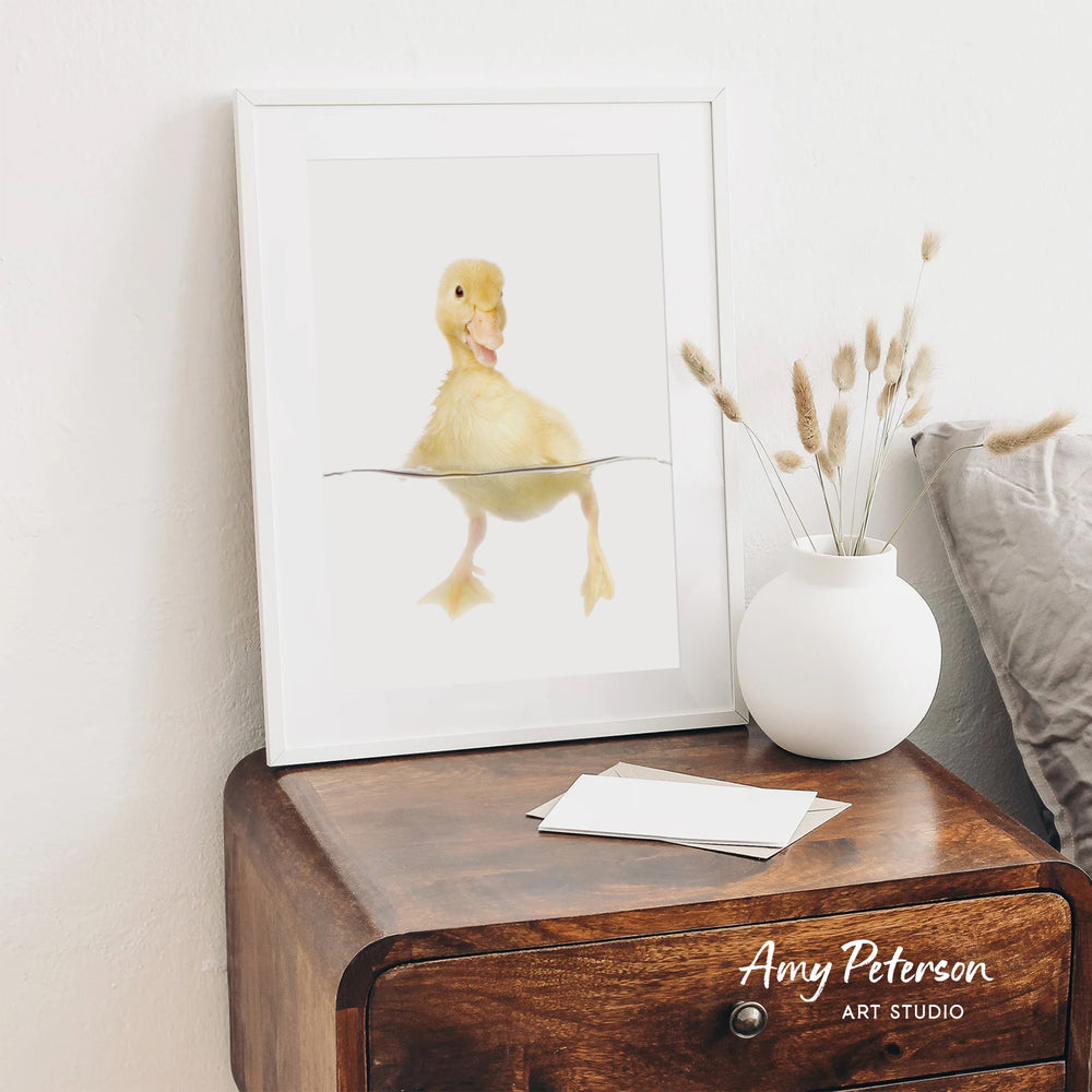 a wooden nightstand with a framed picture of a duck sitting on a branch, a white vase with dried flowers, and some papers on the surface.