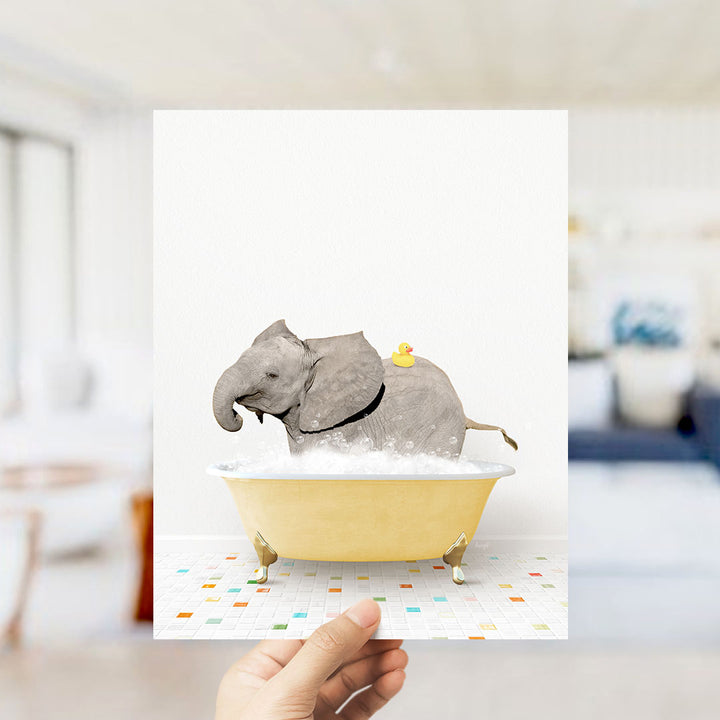 A hand holds up a framed image depicting a gray elephant sitting in a yellow bathtub, with a yellow rubber duck on its back.