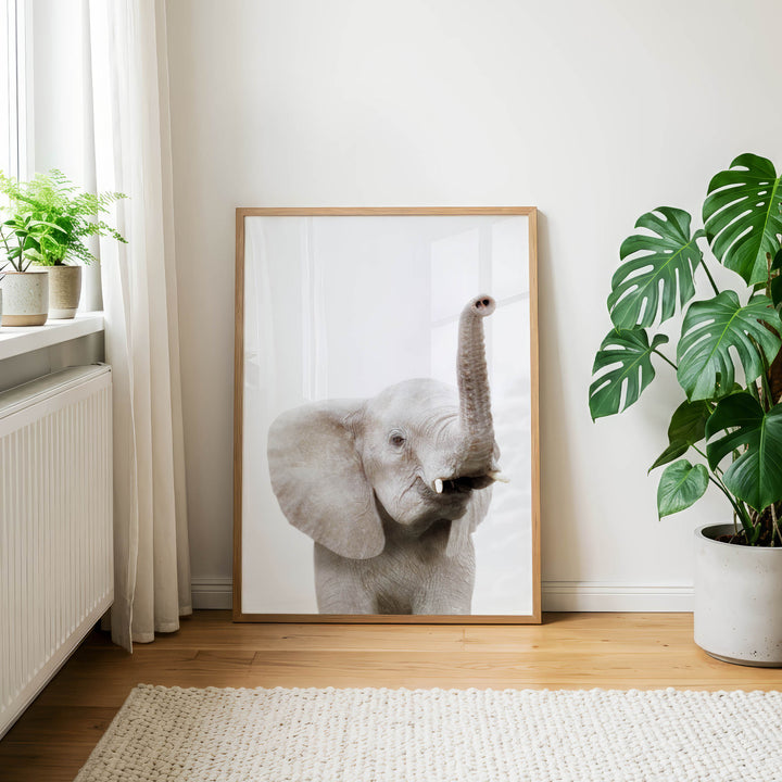 a large elephant standing in a room with a white wall and a window. The elephant is framed and placed on a wooden floor, with a potted plant to the right of the frame.