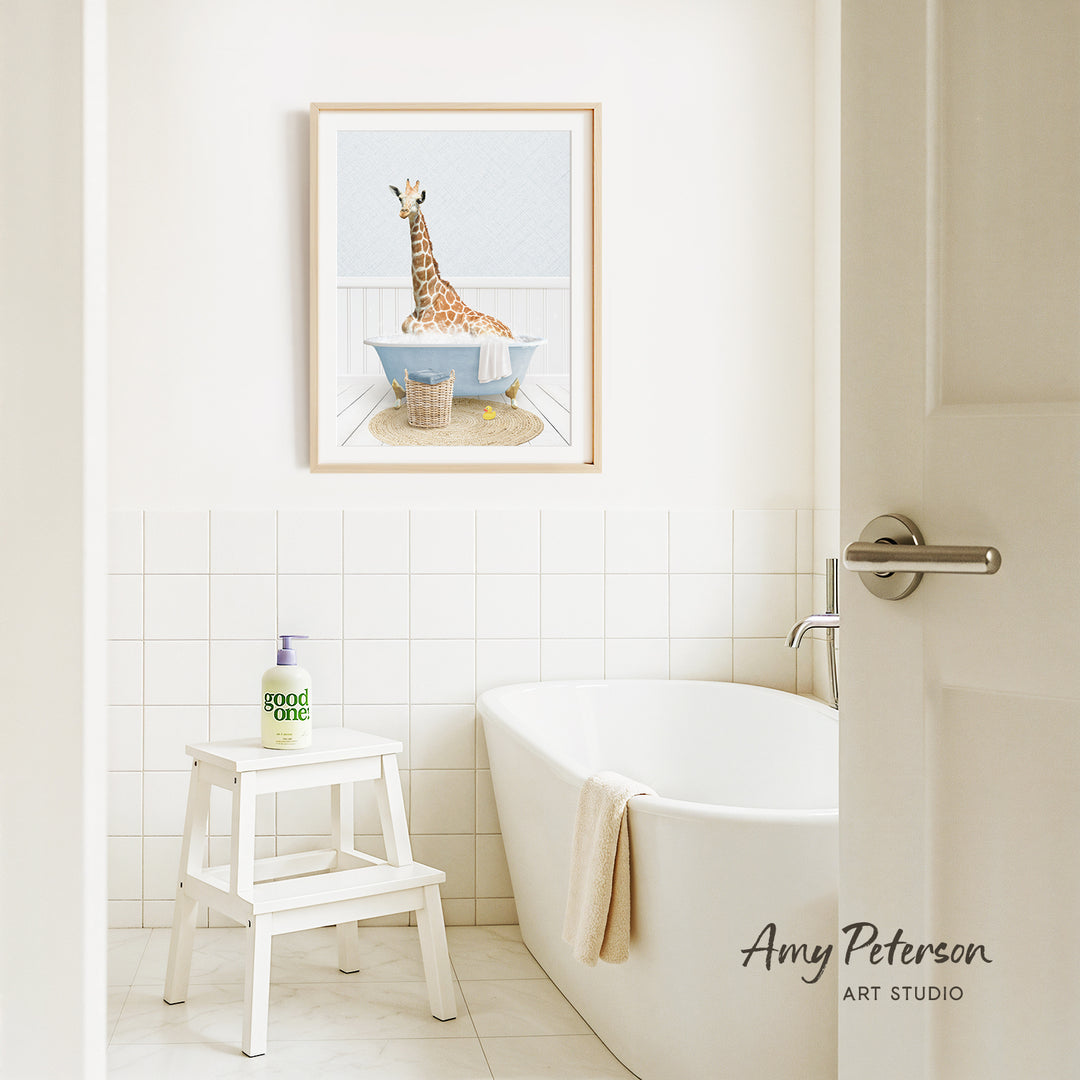 A framed illustration of a giraffe sitting in a bathtub is displayed on the wall above a white bathtub in a bathroom.
