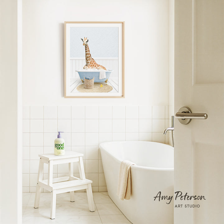 A framed illustration of a giraffe sitting in a bathtub is displayed on the wall above a white bathtub in a bathroom.