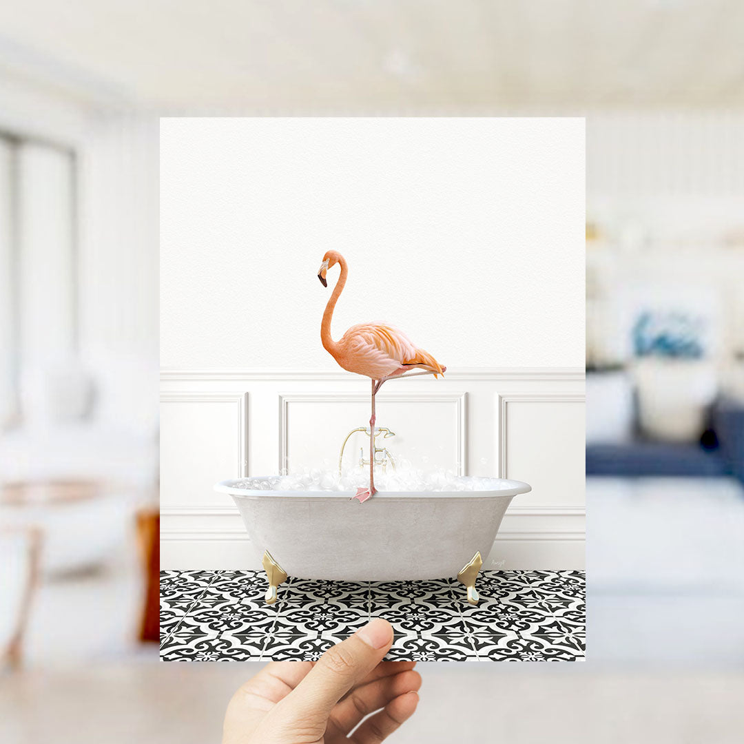 A hand holds up a framed image of a pink flamingo standing on a white bathtub filled with water, set against a white wall with a black and white patterned floor.