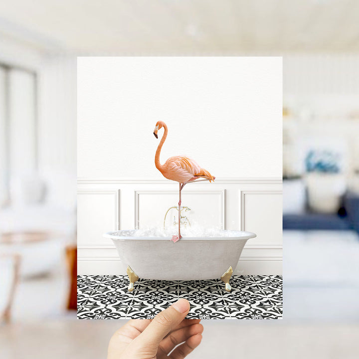 A hand holds up a framed image of a pink flamingo standing on a white bathtub filled with water, set against a white wall with a black and white patterned floor.