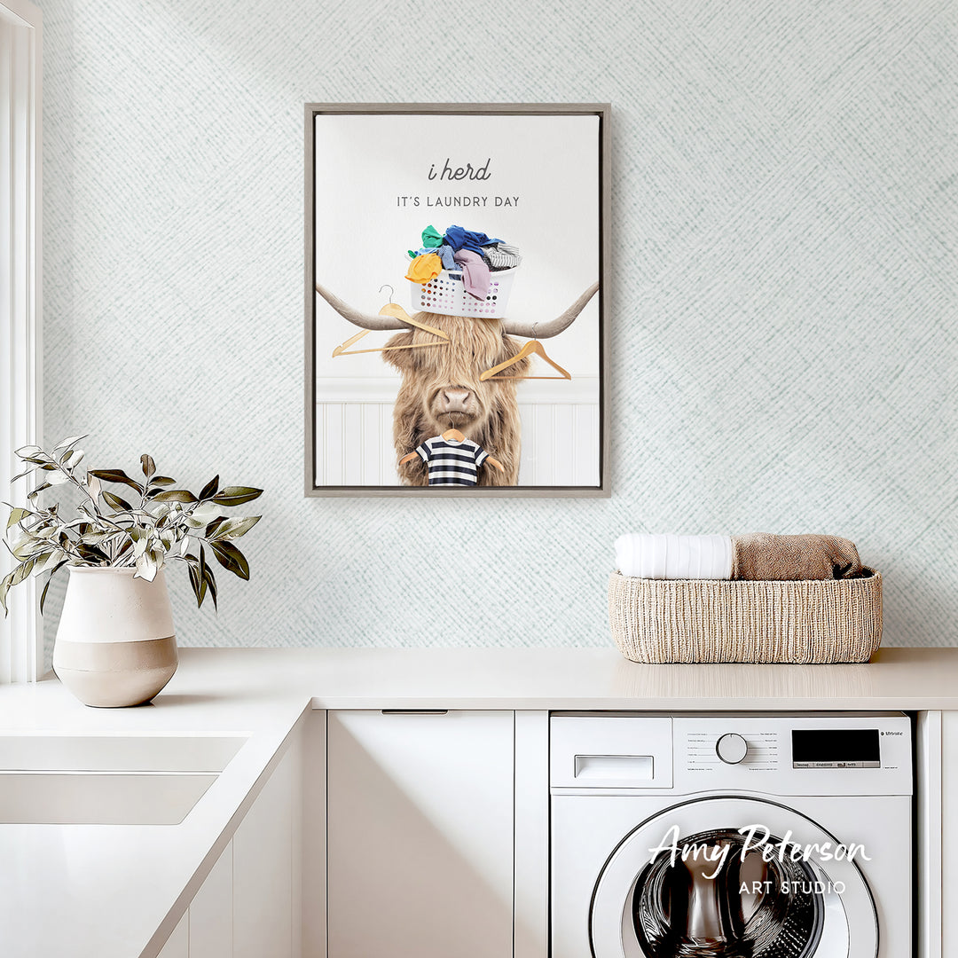 a framed poster of a cow with a basket of laundry hanging on its horns, placed above a white kitchen counter with a potted plant and a basket of towels.