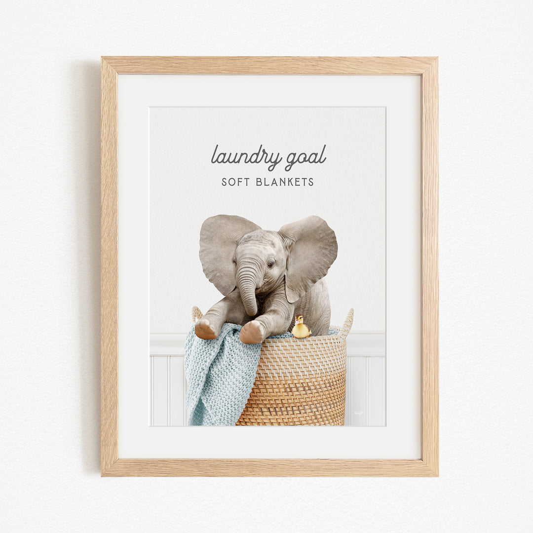 A framed image of a baby elephant sitting in a basket filled with soft blankets, with the text "laundry goal soft blankets" written above the elephant.
