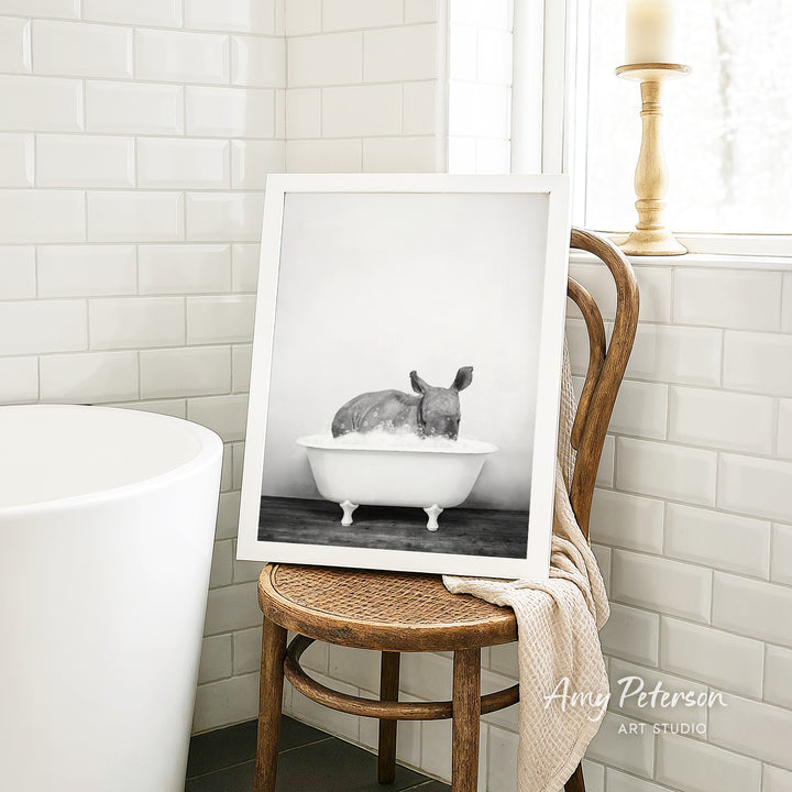 A framed black and white photograph of a rhinoceros sitting in a bathtub, with a towel draped over the back of a wooden chair in a bathroom setting.