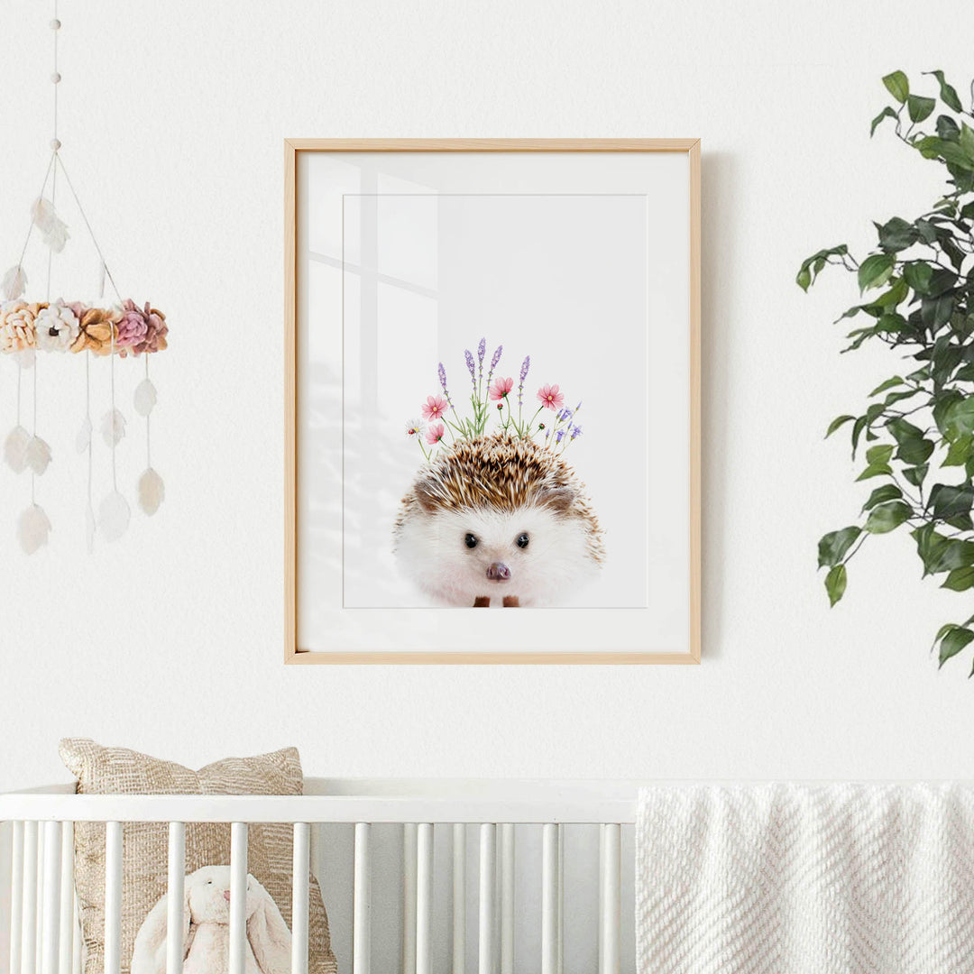 A framed picture of a hedgehog with flowers on its head hangs on a white wall, accompanied by a hanging mobile and a plant.