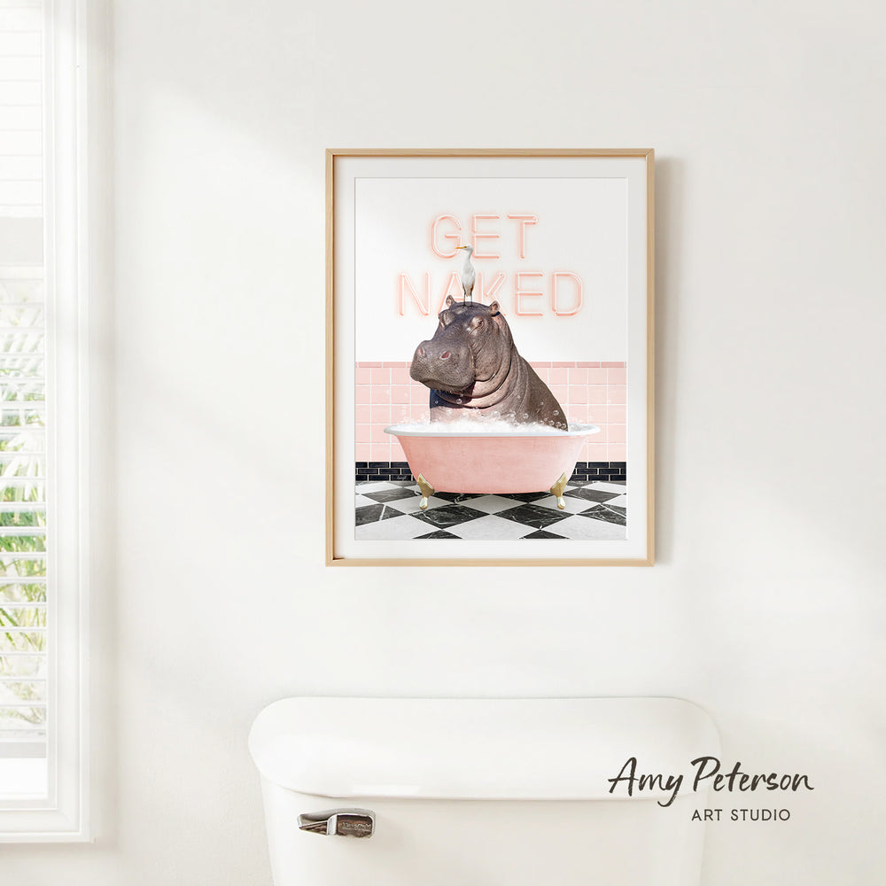 A framed print of a hippopotamus sitting in a pink bathtub with the text "GET NAKED" on it, hanging on a white wall.
