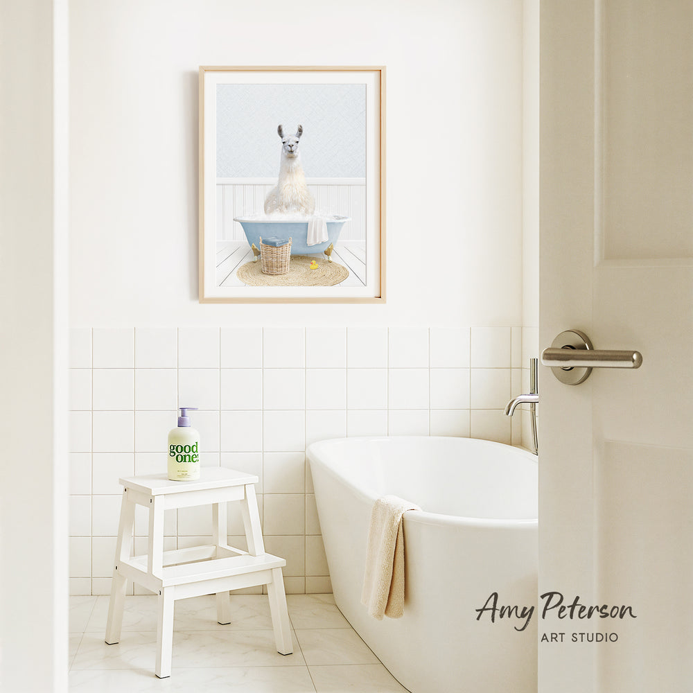 A framed image of a llama sitting in a bathtub is displayed on the wall above a white bathtub in a bathroom.