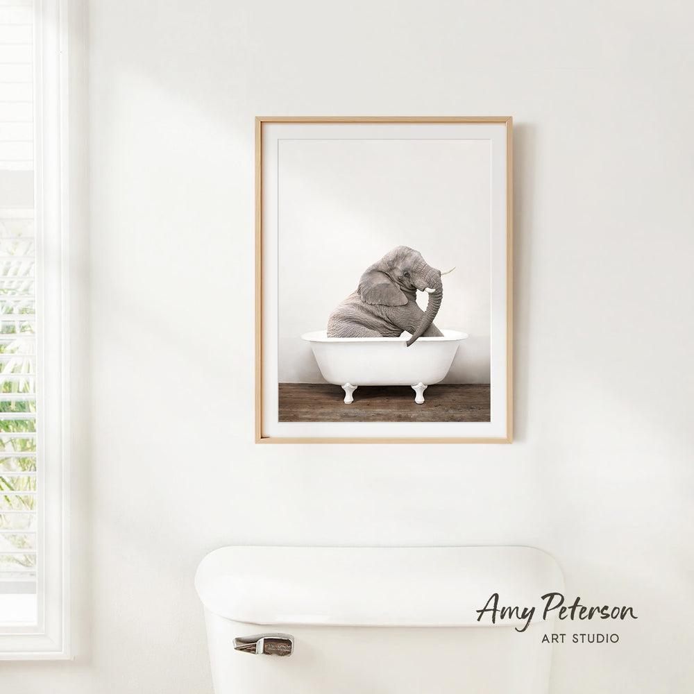 A framed image of an elephant sitting in a bathtub is hanging on a white wall.