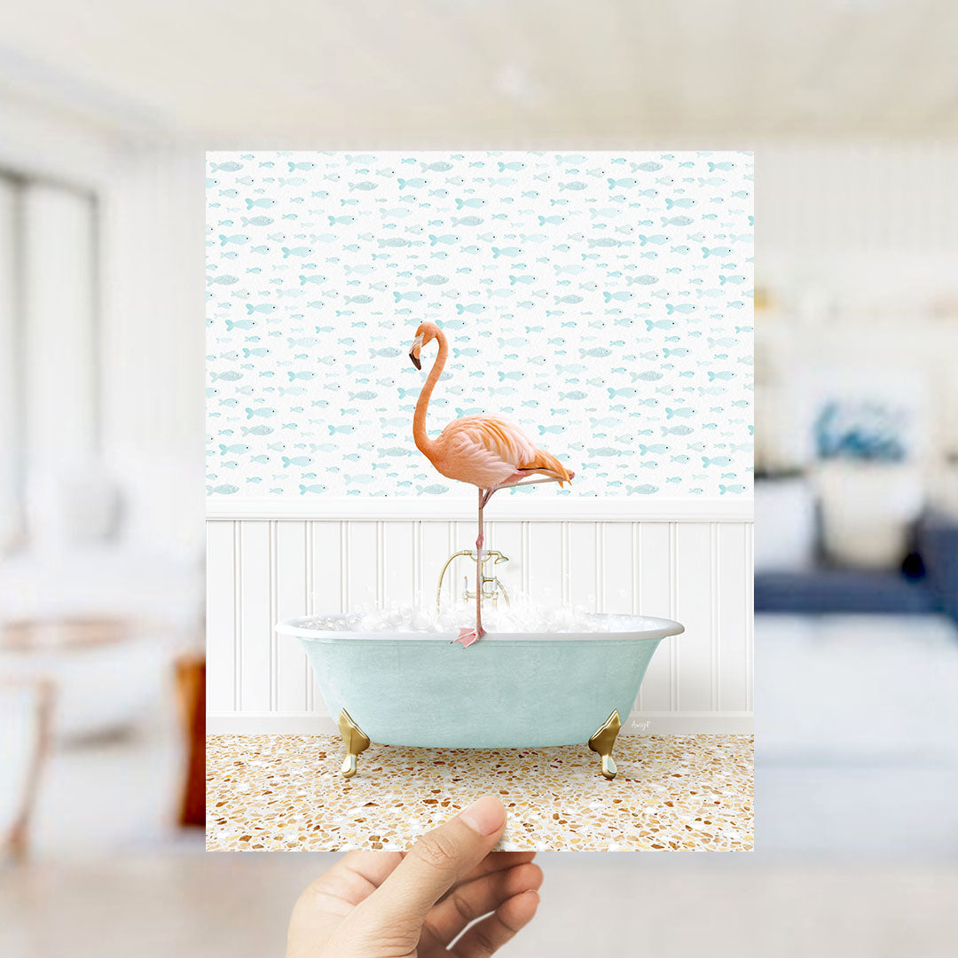 A hand holds up a framed image of a pink flamingo standing in a bathtub filled with bubbles, set against a backdrop of a fish-patterned wall.