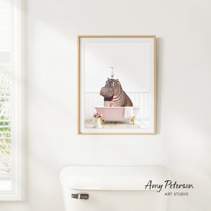 A framed image of a hippopotamus sitting in a pink bathtub, with a window and toilet visible in the background.