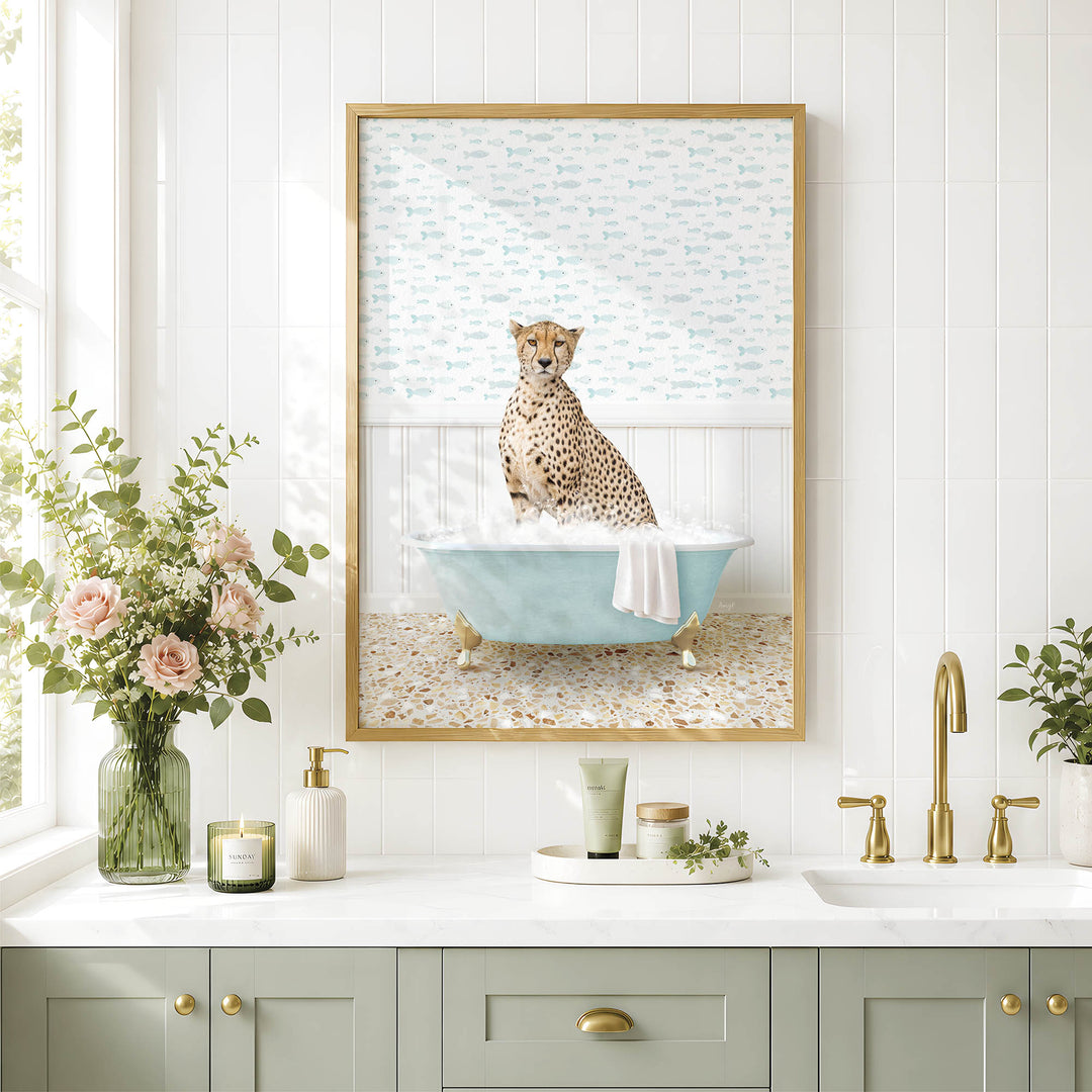 A framed image of a cheetah sitting in a bathtub, surrounded by flowers and candles, is displayed on a white tiled wall above a bathroom counter with a sink and gold faucets.